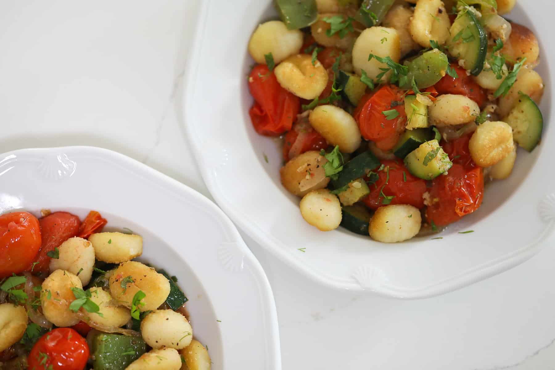 Overhead shot of pan-fried gnocchi with burst cherry tomatoes, zucchini, and green bell peppers in a skillet, garnished with fresh herbs