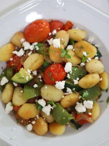 Overhead shot of pan-fried gnocchi with burst cherry tomatoes, zucchini, and green bell peppers in a skillet, garnished with fresh herbs and feta